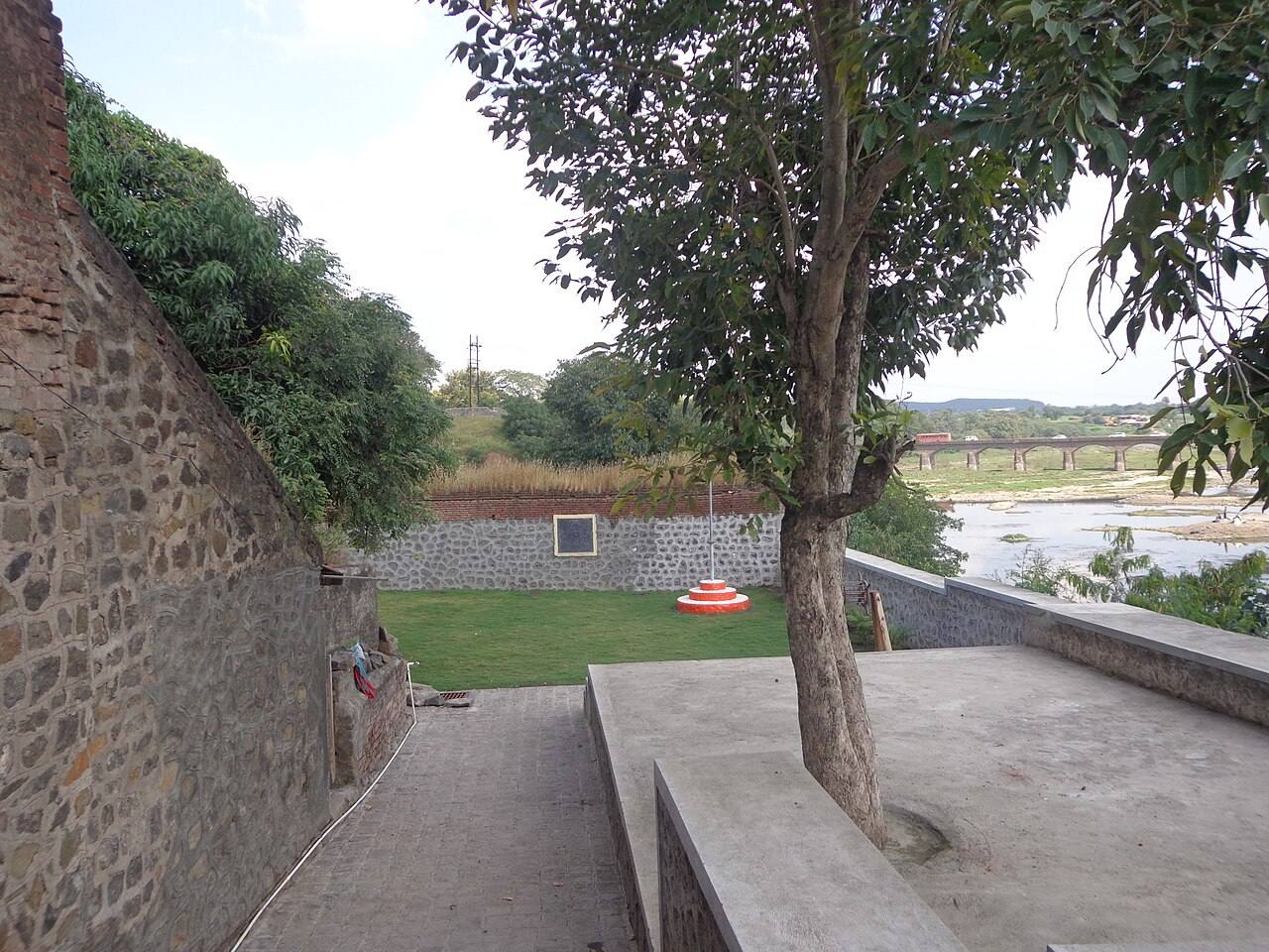 Rajguru Wada memorial at Rajgurunagar with stone walls, a lawn, a flag base and a plaque, overlooking the Bhima river and a bridge