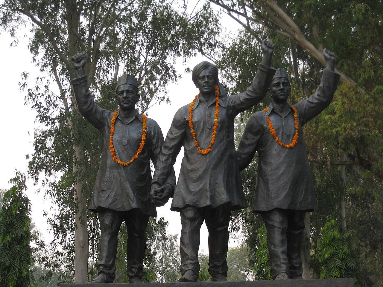 Garlanded bronze statues of Bhagat Singh, Rajguru and Sukhdev with raised fists, standing on a plinth among trees
